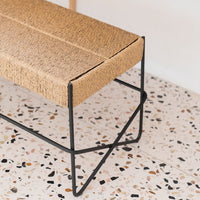 Natural woven beige bench with a minimalist black metal frame, set against a speckled terrazzo floor.