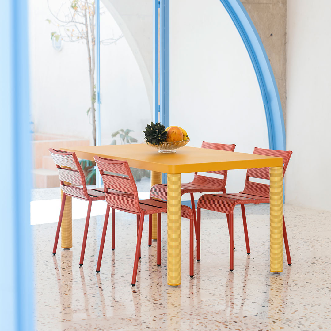 Bright yellow table with red chairs, set against a minimalist backdrop featuring blue accents and a textured floor.