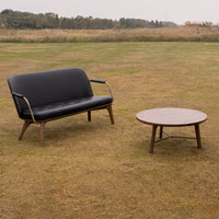 Black two-seater sofa with wooden legs and round brown coffee table, set against a grassy outdoor background.