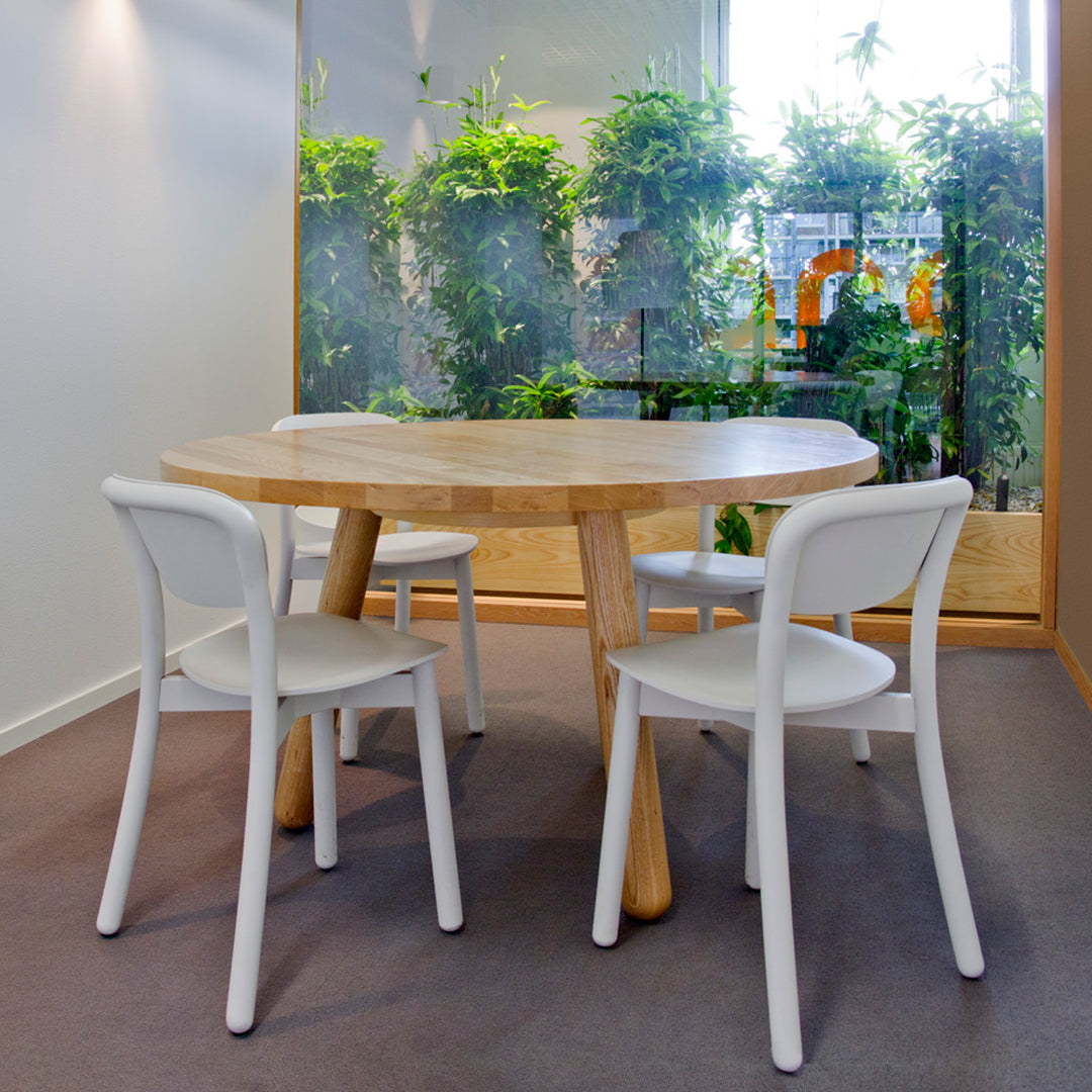 Light wood round table with natural finish and white chairs against a green plant backdrop.