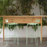 Mint green metal bar stools beside a light wood table, against a backdrop of lush green plants.