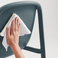 Hand cleaning a sleek, dark teal chair with a white cloth against a minimalist background.