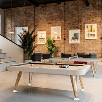 Modern white pool table with wooden legs, colorful balls on the surface, and a stylish industrial backdrop.