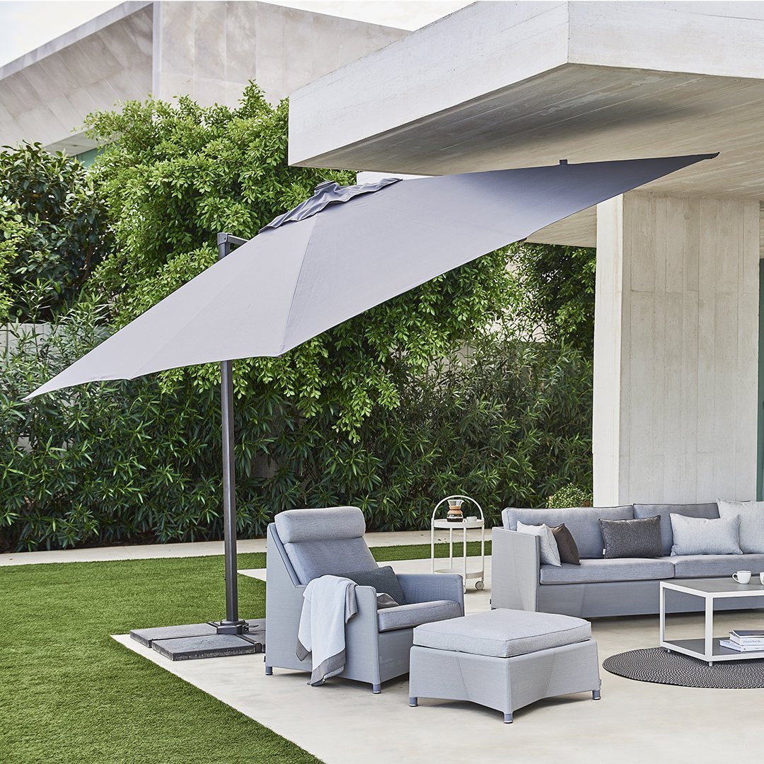 Gray outdoor umbrella, soft gray seating, and patterned throw pillows against lush green foliage and concrete backdrop.