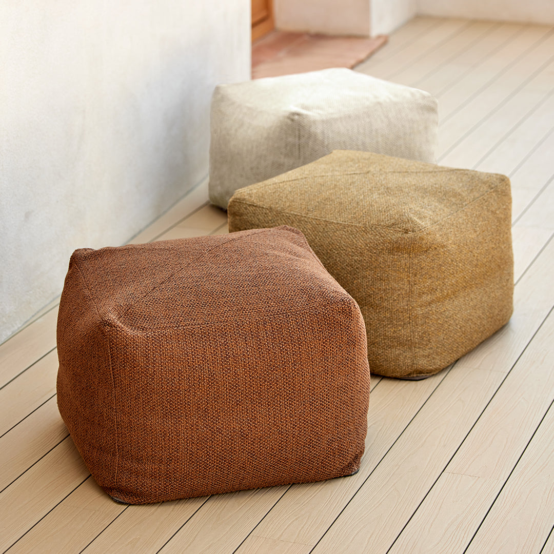 Three cozy poufs in earthy tones: rust, cream, and tan, featuring a textured fabric design on wooden flooring.