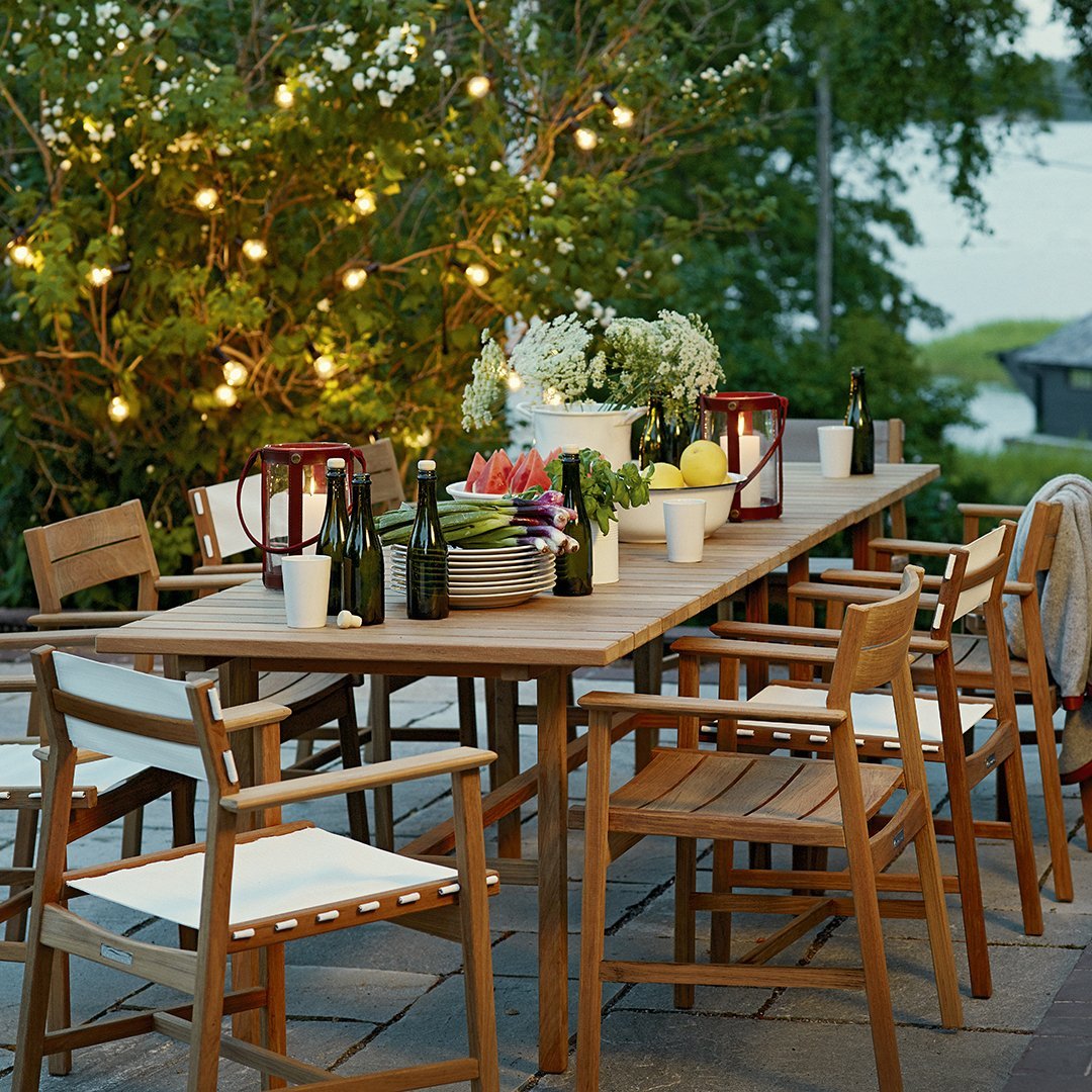 Wooden outdoor dining table with white cushions, colorful floral centerpieces, and ambient warm string lights.
