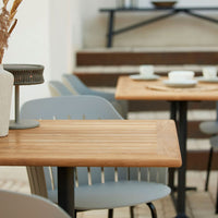 Light wooden table with slatted surface, accompanied by soft gray chairs and a neutral vase, in a bright outdoor setting.