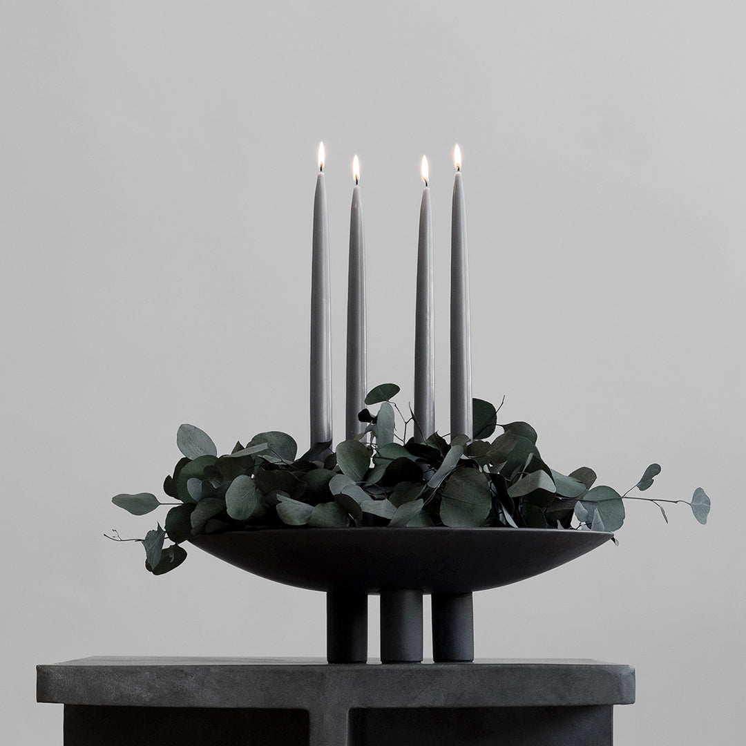 "Elegant black candle holder with three slender candles and green eucalyptus accents, set against a light gray background."