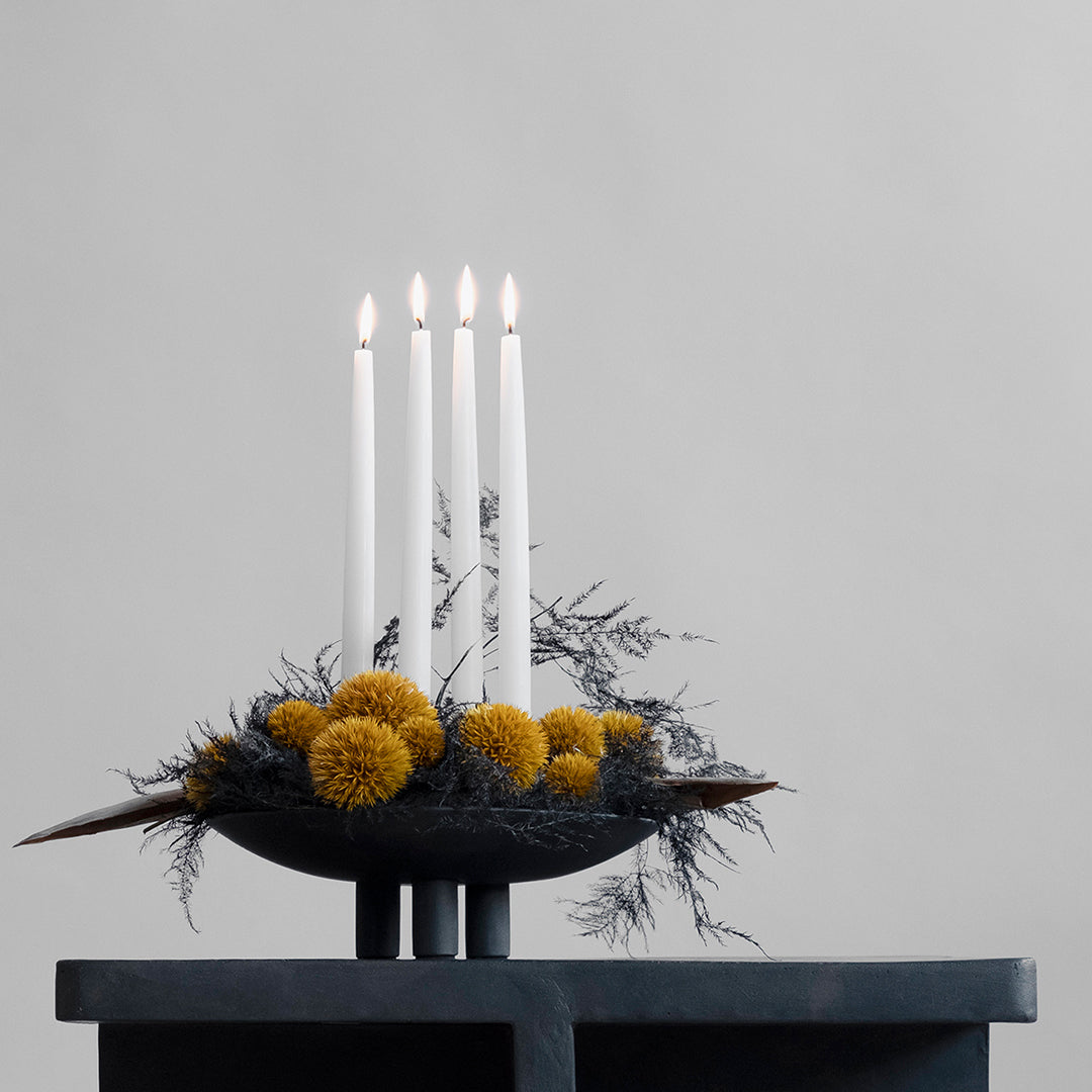 "Black candleholder with four white candles and yellow flowers, set against a soft gray background."
