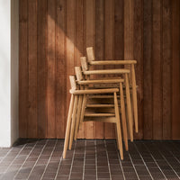 Stacked light brown wooden chairs against a warm, wooden paneled wall, with dark tiled flooring.