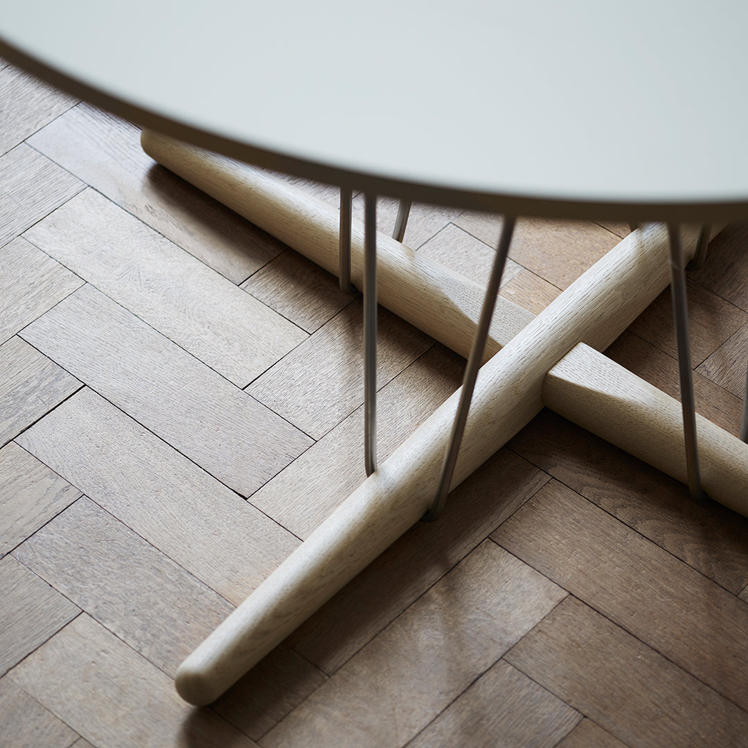 White round table top with natural wood cross legs on a light wooden floor. Minimalist design with soft color tones.