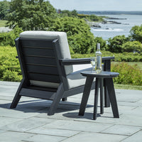 Modern black outdoor chair with gray cushion, paired with a matching black side table, set against a lush green landscape.