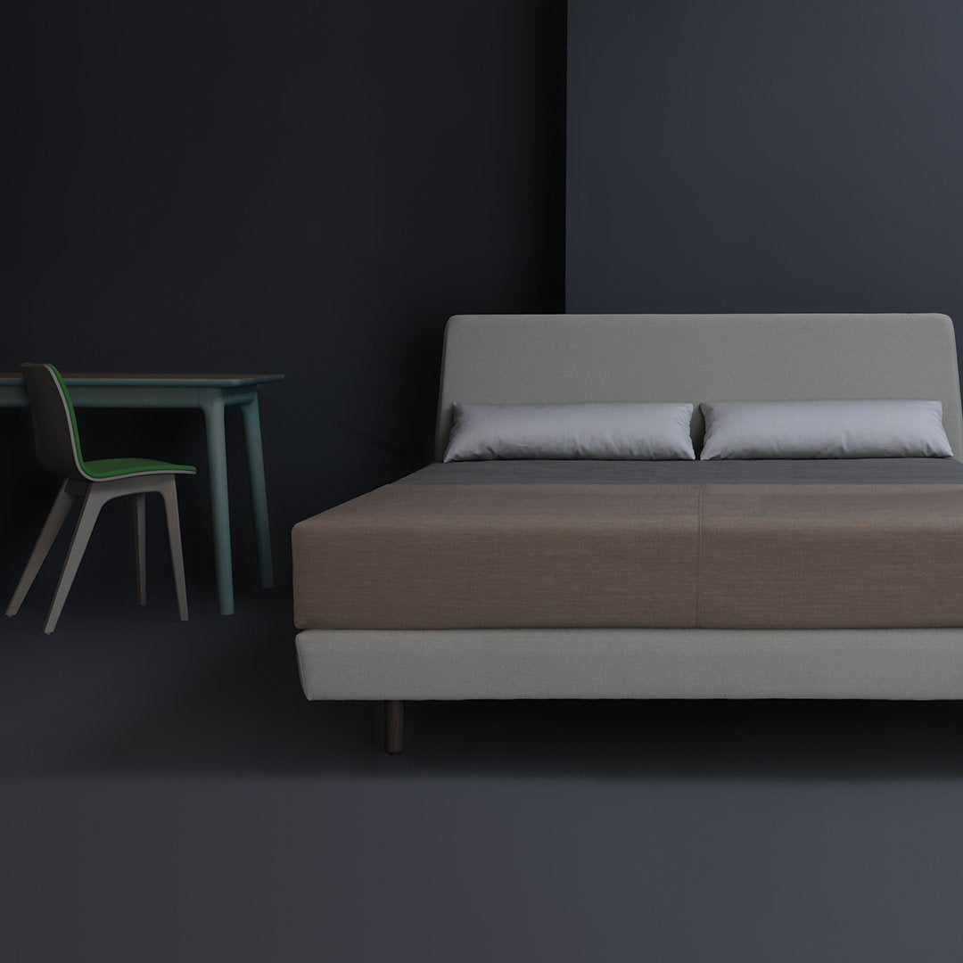 Modern bed in beige and brown with silver pillows, complemented by a minimalist green chair against a dark background.