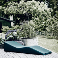 Teal lounge chair with smooth, curved edges against a lush green garden backdrop. Modern, minimalist design.