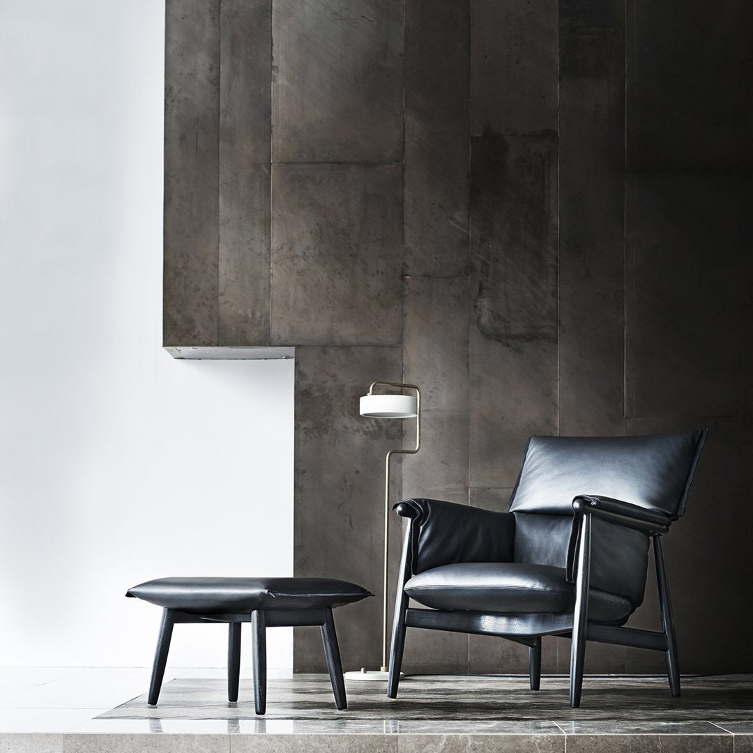 Black leather armchair with sleek lines and matching ottoman, set against a textured gray wall. Minimalist design.