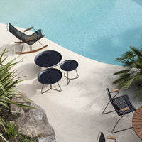 Black circular side tables and striped chairs beside a blue pool with sandy beige surroundings and tropical plants.