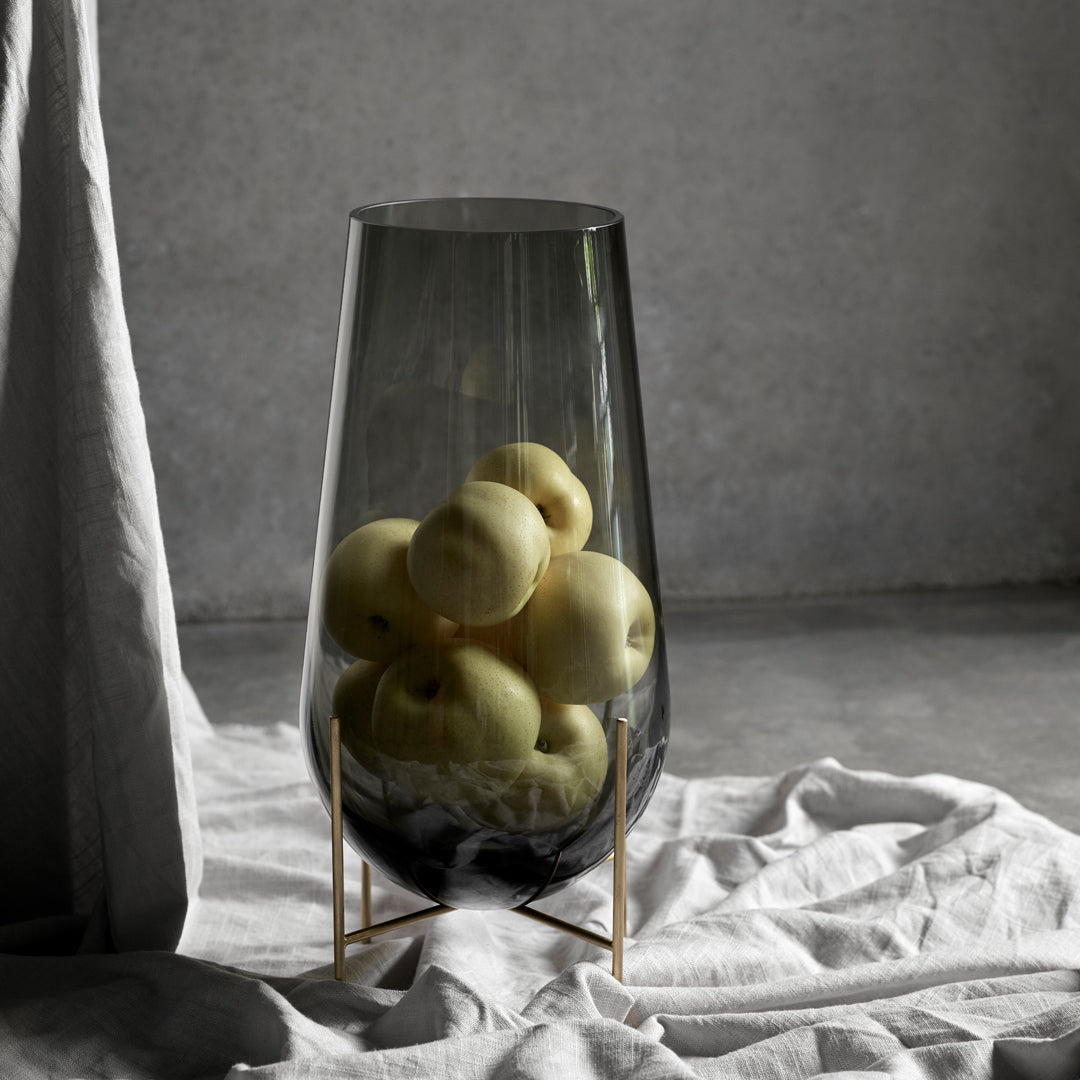 Smoky glass vase with sleek curves, filled with yellow apples, set against soft gray and white textures.