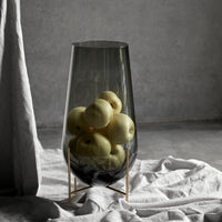 Smoky glass vase with sleek curves, filled with yellow apples, set against soft gray and white textures.