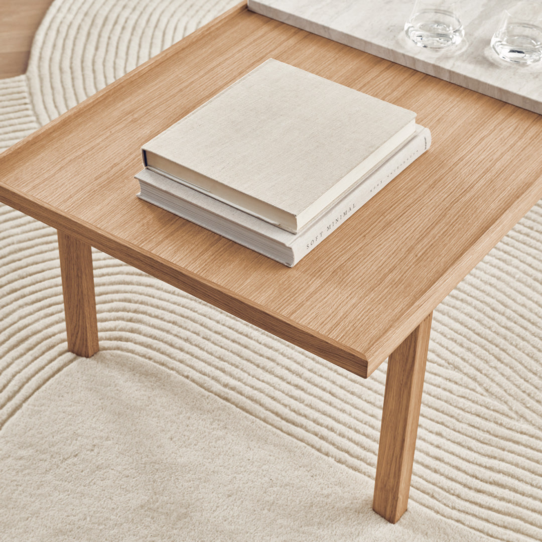 Light wood table with a smooth surface, featuring stacked white books and a neutral, textured rug underneath.