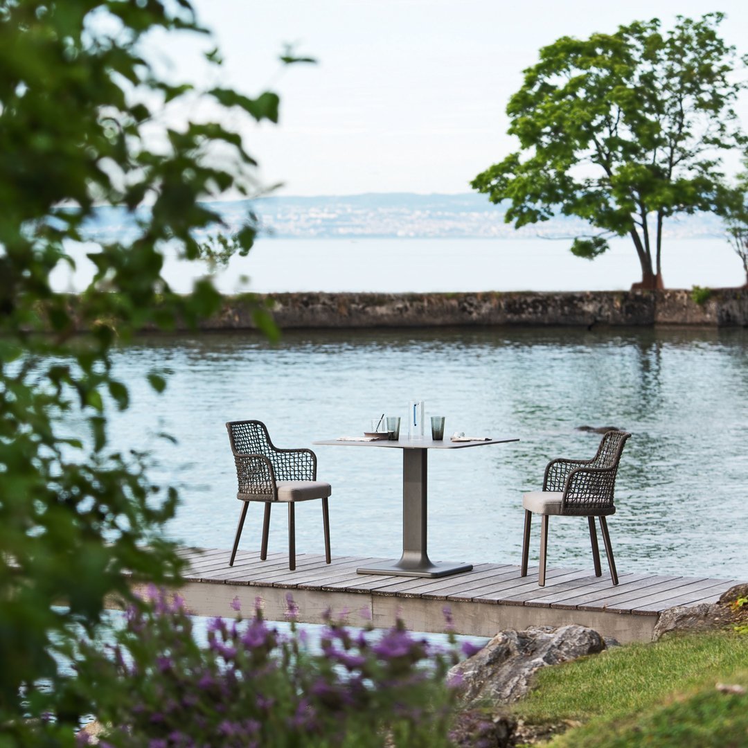 "Two grey wicker chairs and a round table on a wooden deck by a serene blue lake, surrounded by green foliage."