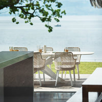 "Neutral-toned outdoor dining set featuring woven wicker chairs and a sleek white table against a serene lake backdrop."