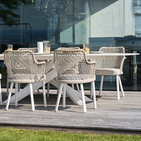 Light beige woven chairs with white legs around a modern white table on a wooden deck, set against a glass backdrop.