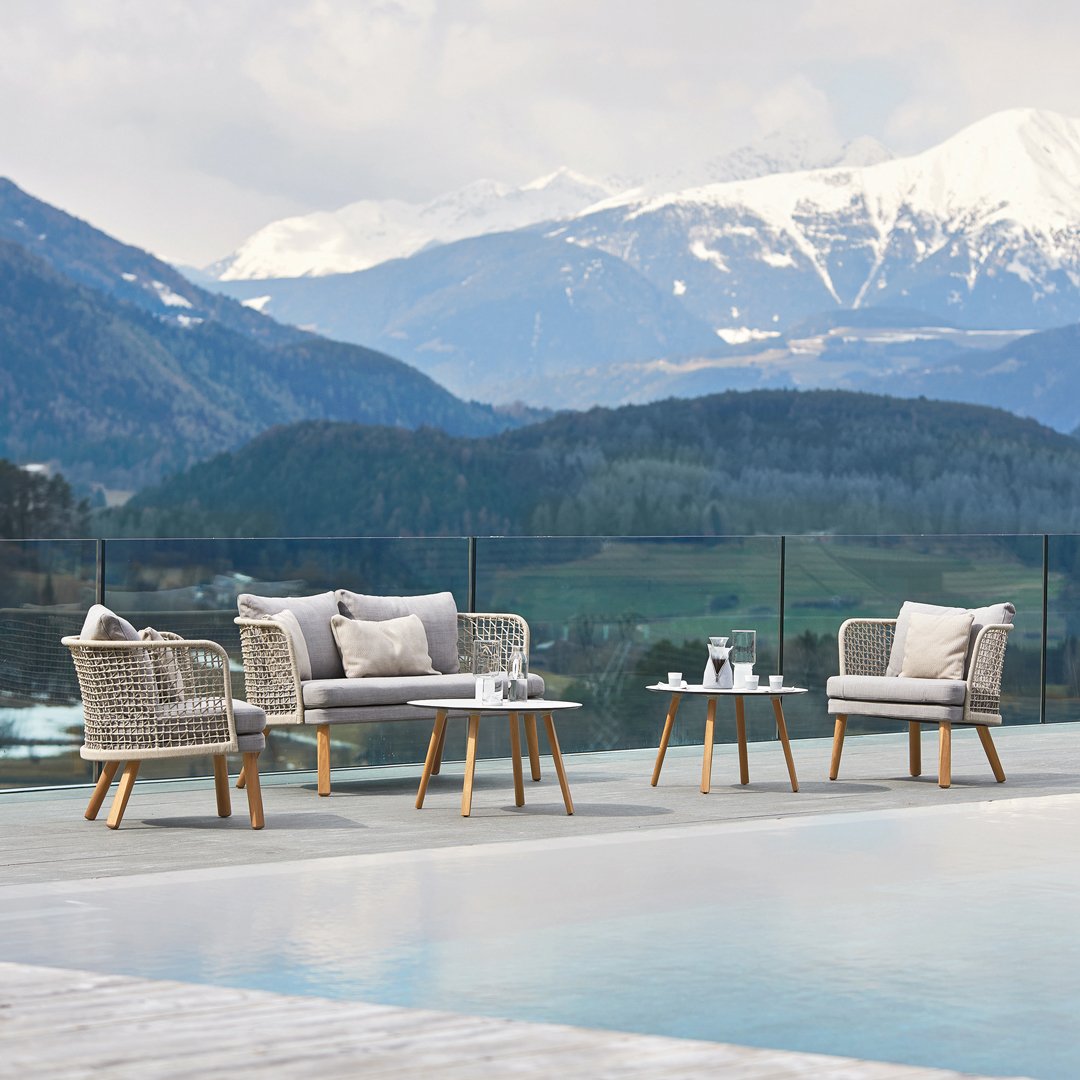 "Light grey woven outdoor furniture with soft white cushions, set against a scenic mountain backdrop."