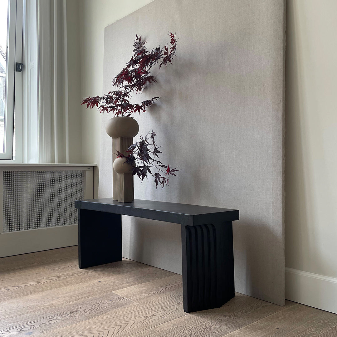 "Black wooden bench with ribbed edges, set against a light gray background and accented by a brown vase and red foliage."