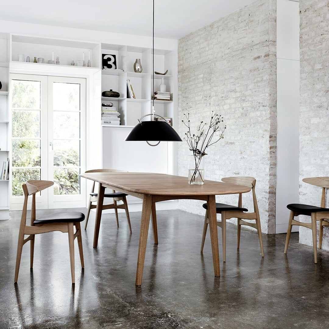 Modern wooden dining table with light oak finish and black patterned chairs, set against a neutral-toned backdrop.
