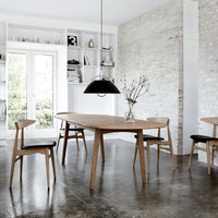 Modern wooden dining table with light oak finish and black patterned chairs, set against a neutral-toned backdrop.