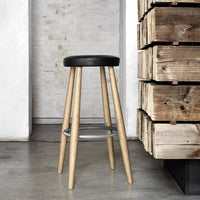 Black round seat with natural wood legs and a silver footrest, against a textured concrete background.