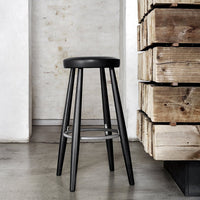 Black wooden stool with a round seat and sleek legs, featuring a minimalist design against a textured backdrop.