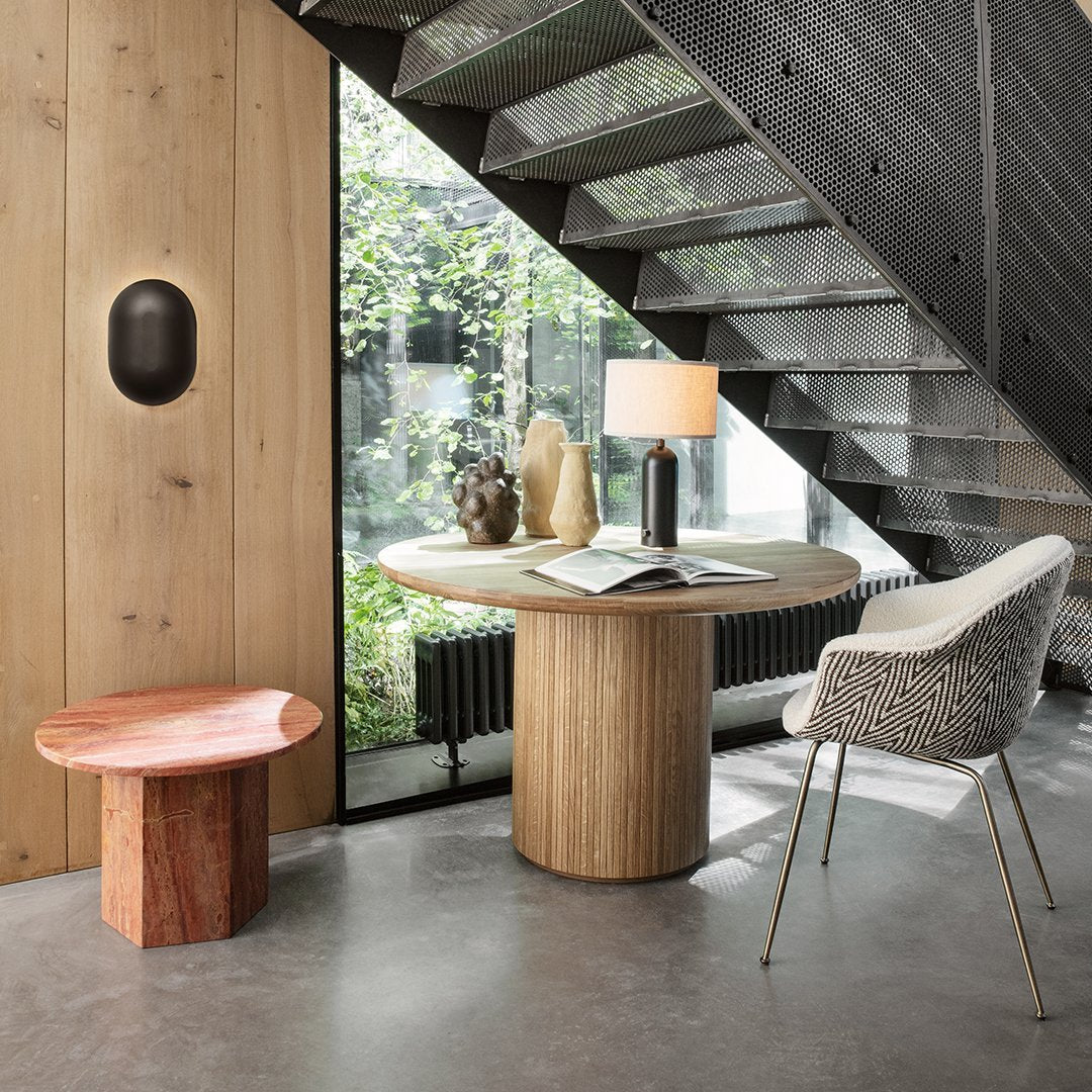 Modern interior featuring a round wooden table in light natural tones, paired with a red accent stool and a white textured chair.