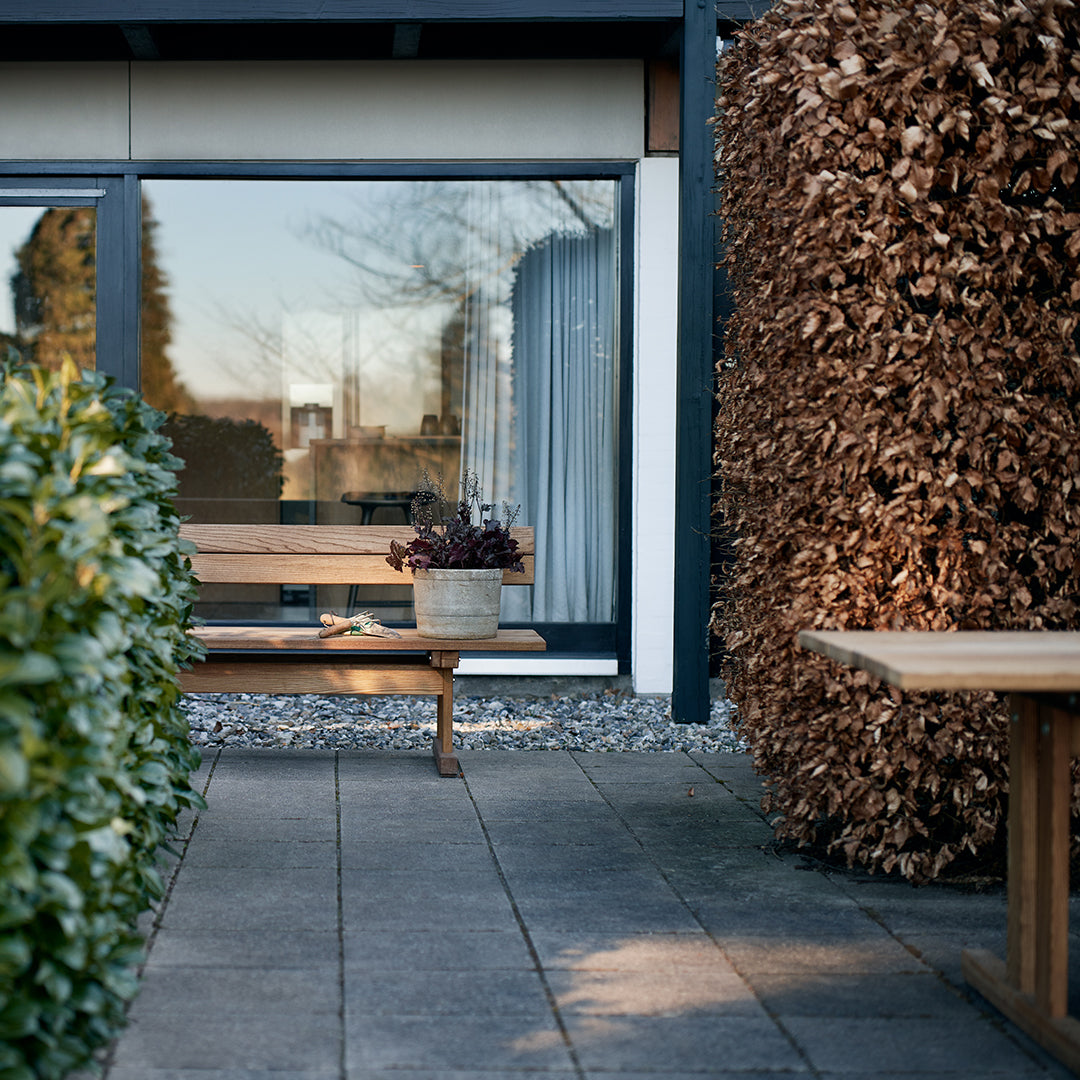 "Green foliage and brown textured hedge frame a modern patio with a light wooden table and neutral exterior colors."