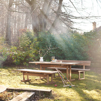 Natural wood dining table and benches in a sunlit garden with lush green foliage and soft sunlight patterns.