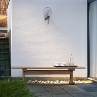 Natural wood bench in warm tones, set against a textured white wall and complemented by a clear glass lamp.