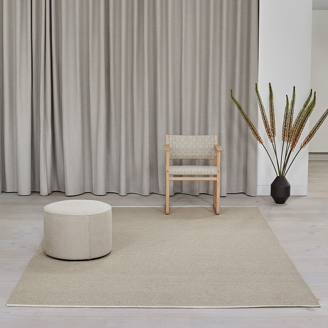 Neutral beige area rug with subtle texture, paired with light wood chair and soft cream pouf in a minimalist setting.