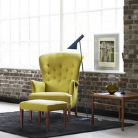Yellow tufted armchair with matching ottoman on a black rug, set against a brick wall and large windows.