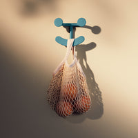 "Light beige wall with a mint green hook holding a white mesh bag, showcasing the warmth of orange fruits inside."