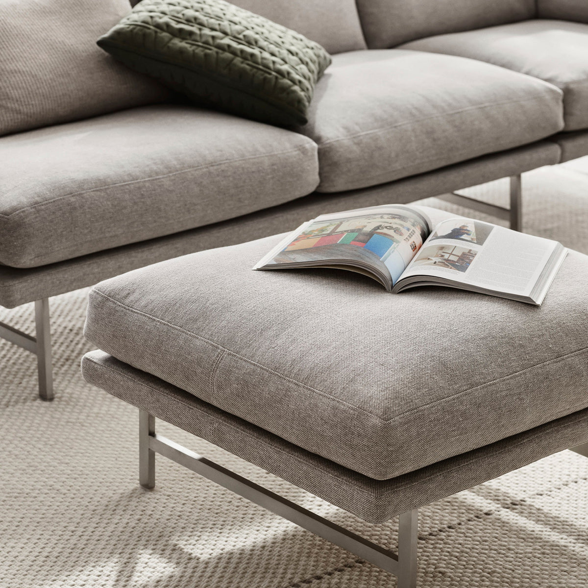 Gray textured sofa with coordinating ottoman and a green knitted pillow, set on a woven beige rug.