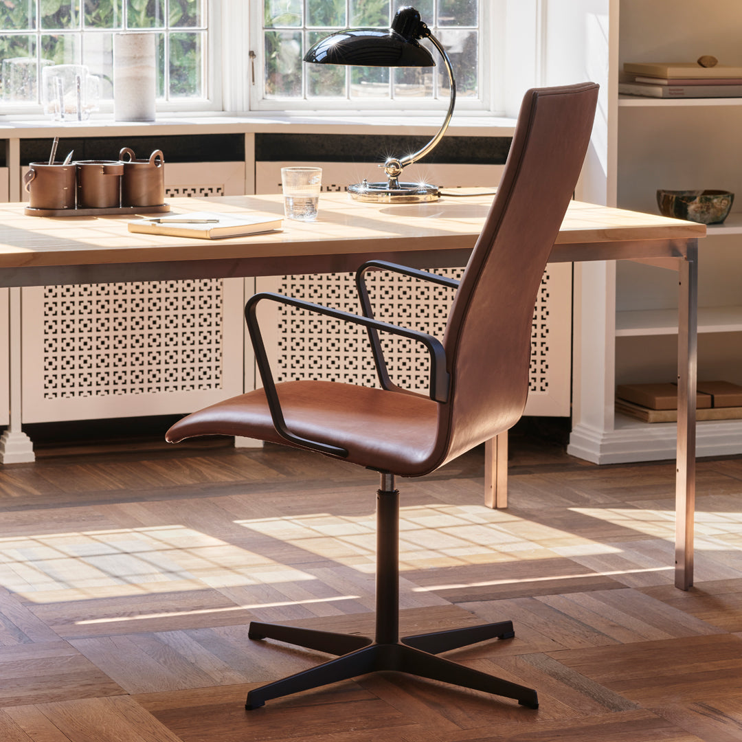 Brown leather office chair with sleek black metal frame, set against a modern interior with warm wooden flooring.