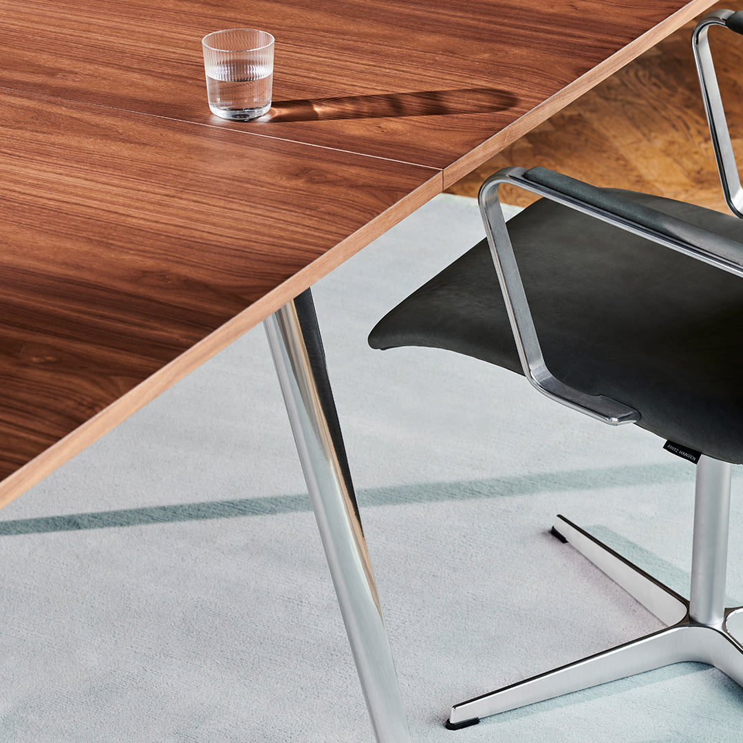 Modern walnut table with polished chrome legs and a sleek black chair on a light gray carpet.