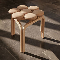 Beige upholstered stool with a geometric pattern, supported by a light wood base, set against a dark tile floor.