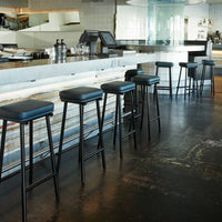 Black leather bar stools with sleek metal legs against a light wood and white bar backdrop. Modern and minimalist design.