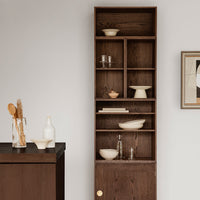 Tall wood shelving unit in rich brown, featuring minimalist white ceramic dishes and glassware arranged in geometric patterns.