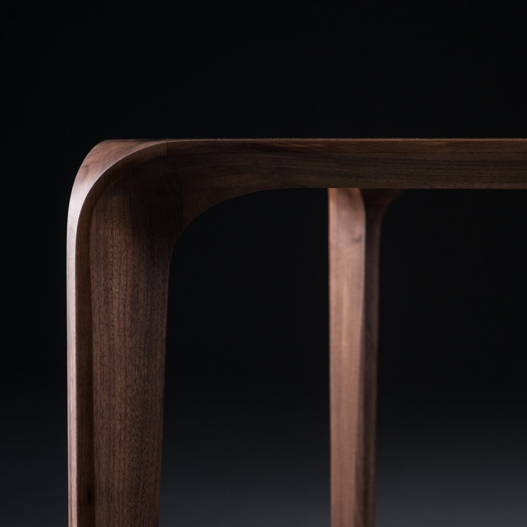 Sleek walnut wood table with smooth curves and rich dark brown tones against a black background.