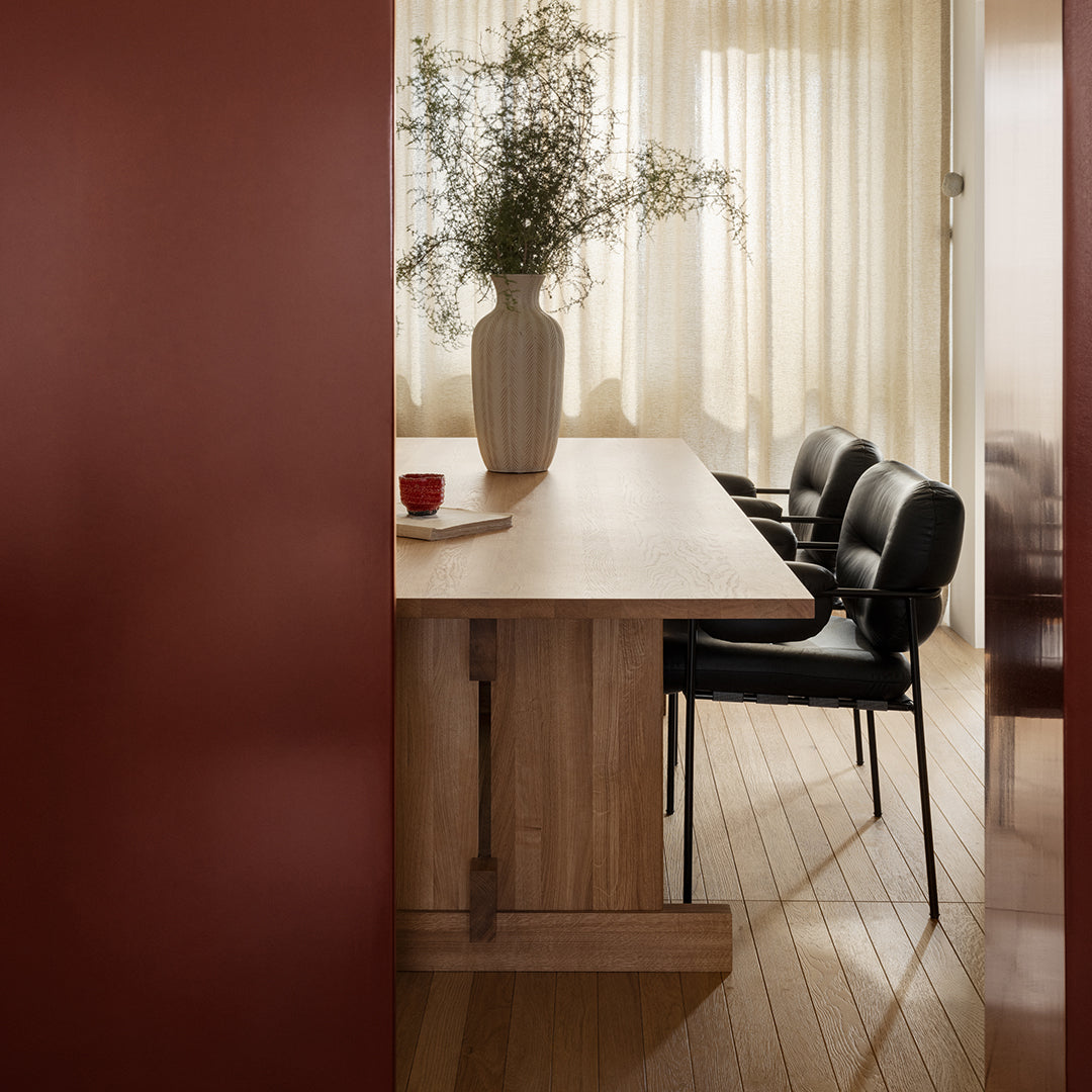 Modern wooden dining table with black chairs, warm wood flooring, and a rich burgundy wall. Neutral curtains and vase accents.