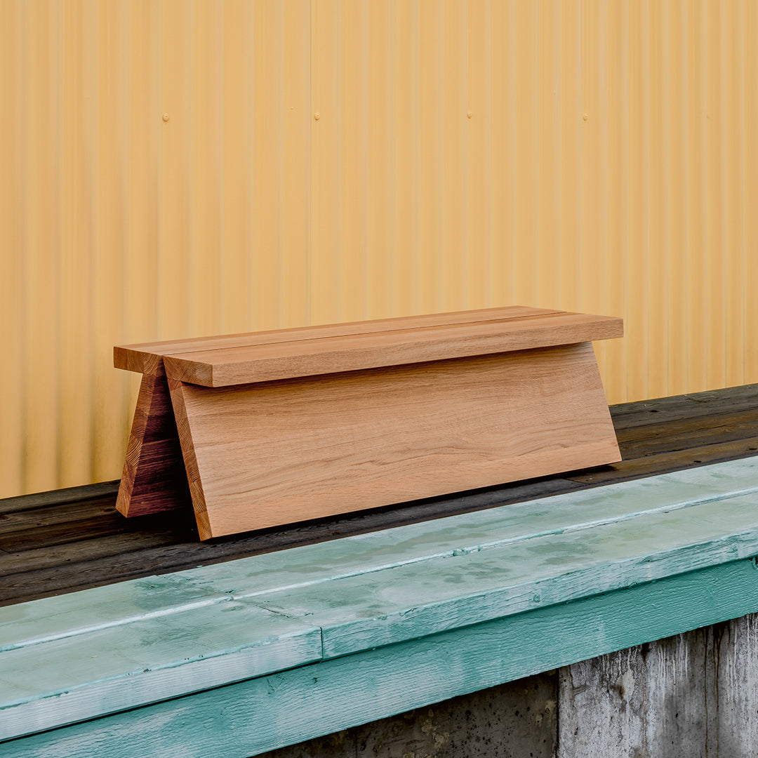 Natural wooden bench with a smooth finish, set against a vibrant yellow corrugated background and rustic blue-green surface.