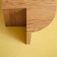 Wooden coffee table with a circular top and contrasting square base on a yellow background. Natural oak finish.