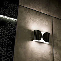 Modern black and silver wall lamp with smooth curves against a textured gray wall featuring a hexagonal pattern.